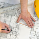 A person is studying blueprints while holding a pencil, with a yellow construction helmet and measuring tools nearby.