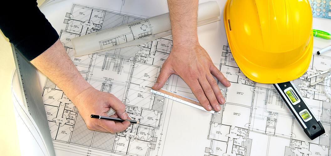 A person is studying blueprints while holding a pencil, with a yellow construction helmet and measuring tools nearby.